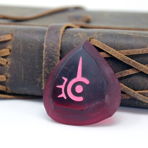 May include: A red, teardrop-shaped guitar pick with a pink symbol on it. The symbol is a stylized eye with a spike coming out of the top. The pick is sitting on a brown leather journal with a braided strap.