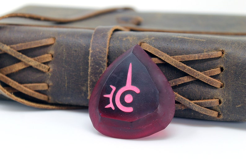 May include: A red, teardrop-shaped guitar pick with a pink symbol on it. The symbol is a stylized eye with a spike coming out of the top. The pick is sitting on a brown leather journal with a braided strap.