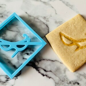 May include: A blue plastic cookie cutter in the shape of a masquerade mask, next to a cookie with the same design imprinted on it.