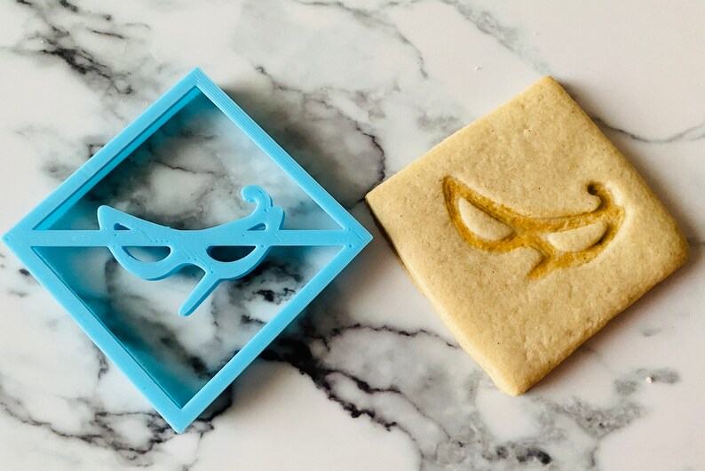 May include: A blue plastic cookie cutter in the shape of a masquerade mask, next to a cookie with the same design imprinted on it.