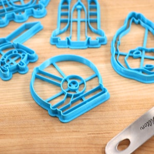 May include: A set of five blue plastic cookie cutters shaped like a spaceship, a helmet, a droid, a dog, and a spaceship. The cutters are on a wooden surface.