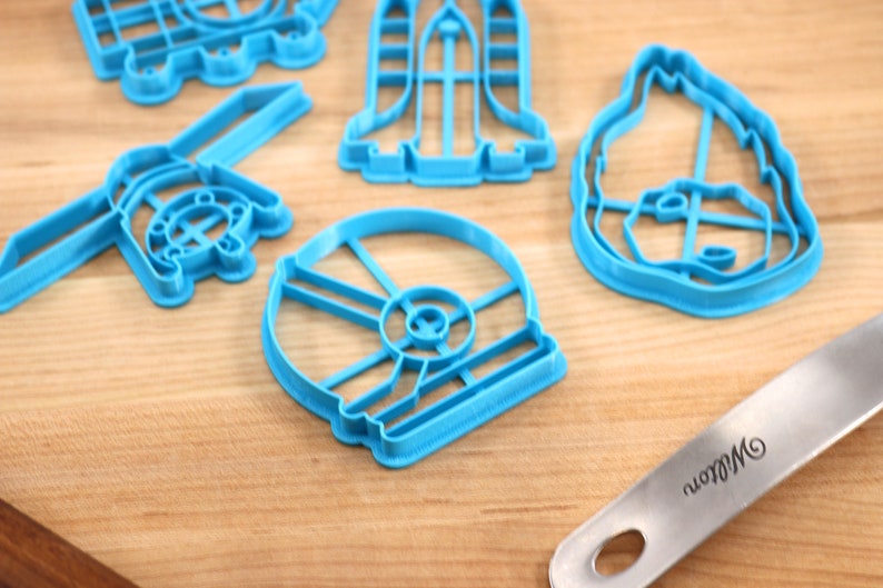 May include: A set of five blue plastic cookie cutters shaped like a spaceship, a helmet, a droid, a dog, and a spaceship. The cutters are on a wooden surface.