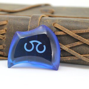 May include: A blue guitar pick with a white symbol on it. The pick is sitting on a brown leather journal.