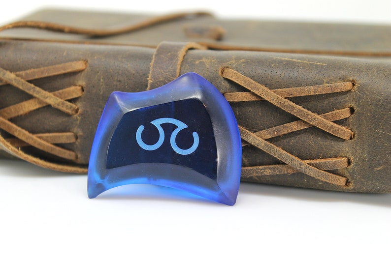 May include: A blue guitar pick with a white symbol on it. The pick is sitting on a brown leather journal.