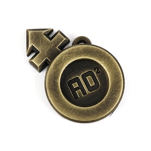 May include: A gold-colored metal charm with a black circle in the center. The circle has the text "AD2" inside. The charm is attached to a small arrow-shaped piece of metal.