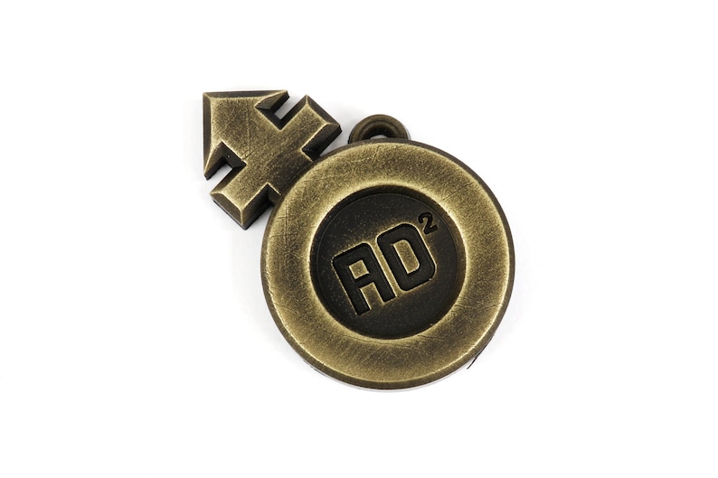 May include: A gold-colored metal charm with a black circle in the center. The circle has the text "AD2" inside. The charm is attached to a small arrow-shaped piece of metal.