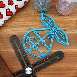 May include: A blue plastic baby-themed cookie cutter with a bow design, alongside a black adjustable angle ruler, a rolling pin, a blue spatula, and a heart-patterned fabric. Sprinkles are in clear containers. Baking and crafting supplies.