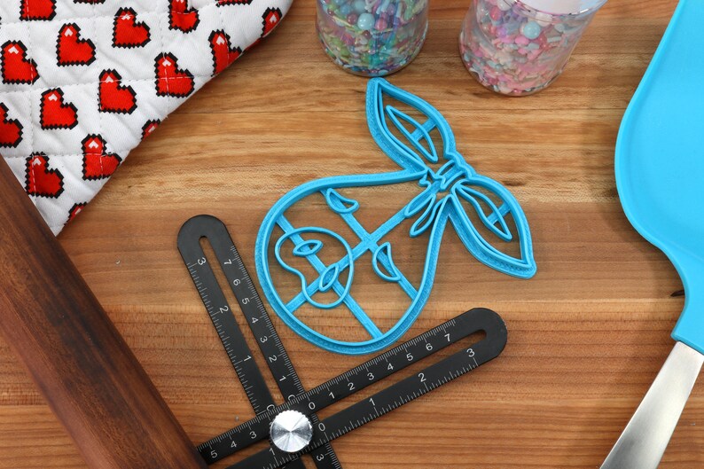 May include: A blue plastic baby-themed cookie cutter with a bow design, alongside a black adjustable angle ruler, a rolling pin, a blue spatula, and a heart-patterned fabric. Sprinkles are in clear containers. Baking and crafting supplies.