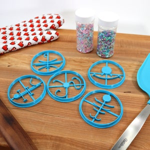 Op de afbeelding: Vijf blauwe plastic koekjesvormen in verschillende vormen op een houten snijplank. Er zijn twee potten met kleurrijke sprinkles en een blauwe spatel.