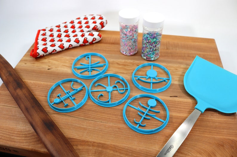 Op de afbeelding: Vijf blauwe plastic koekjesvormen in verschillende vormen op een houten snijplank. Er zijn twee potten met kleurrijke sprinkles en een blauwe spatel.