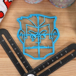 May include: A blue plastic cookie cutter shaped like a person's face with a serious expression. The cutter has a grid pattern on the face and hair.