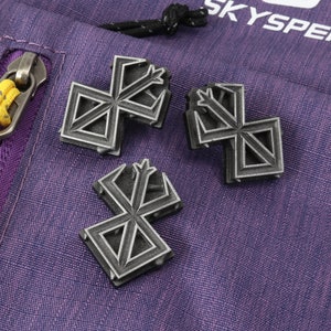 May include: Three silver-coloured metal pins with a geometric design. The pins are on a purple fabric background.