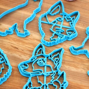 May include: Five blue plastic cookie cutters shaped like dogs. The cutters are on a wooden surface.