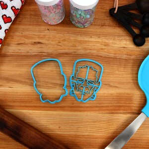 May include: Two blue plastic cookie cutters shaped like Frankenstein's face. One cutter is a detailed outline of the face, while the other is a solid shape.
