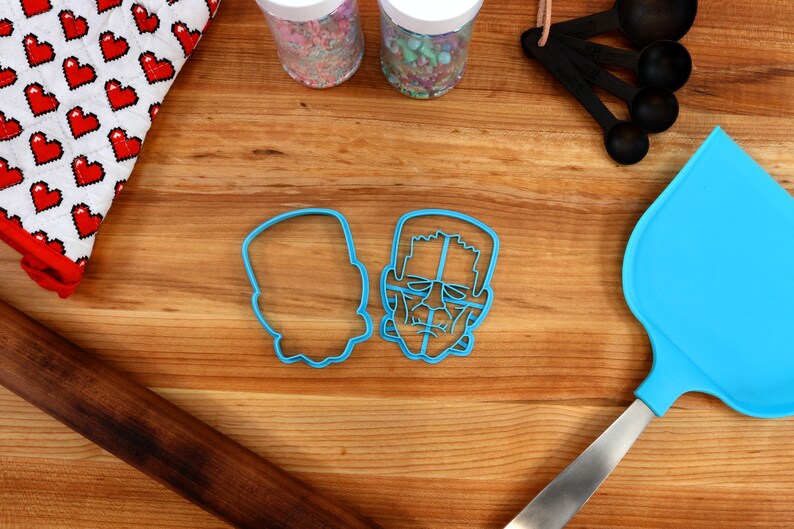 May include: Two blue plastic cookie cutters shaped like Frankenstein's face. One cutter is a detailed outline of the face, while the other is a solid shape.