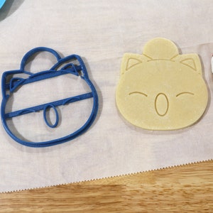 May include: Two cookie cutters, one blue and one beige, shaped like cartoon characters. The blue cutter is of a character with a round head, large eyes, and a small mouth. The beige cutter is of a character with a round head, large eyes, and a small mouth. The cutters are on a white surface.