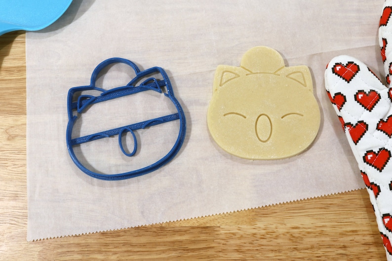 May include: Two cookie cutters, one blue and one beige, shaped like cartoon characters. The blue cutter is of a character with a round head, large eyes, and a small mouth. The beige cutter is of a character with a round head, large eyes, and a small mouth. The cutters are on a white surface.
