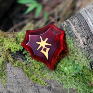 May include: A red, star-shaped resin charm with a gold Chinese character on it. The charm is sitting on a piece of wood covered in moss.