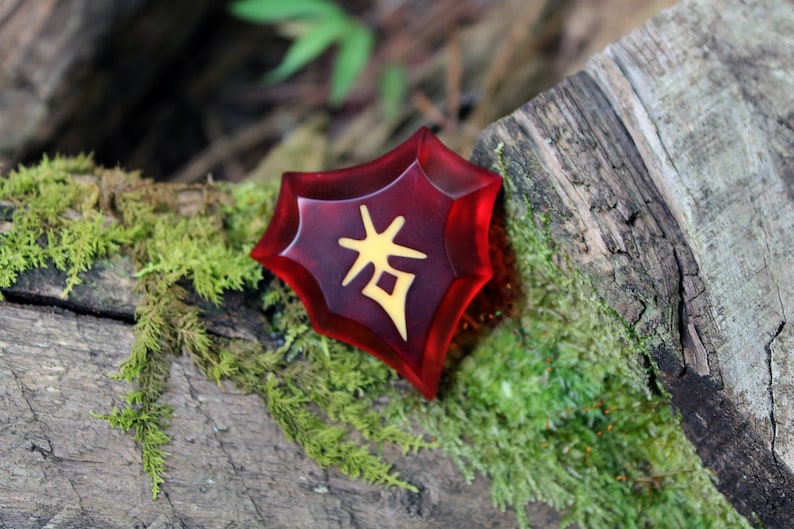 May include: A red, star-shaped resin charm with a gold Chinese character on it. The charm is sitting on a piece of wood covered in moss.