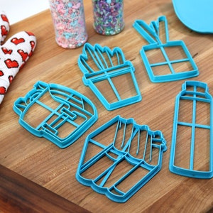 K&ouml;nnte beinhalten: Ein Set mit sechs blauen Plastik-Ausstechern in Form eines Kaktus, einer Geschenkbox, einer Spr&uuml;hflasche, eines Rucksacks, einer Portion Pommes und eines Blumentopfs. Die Ausstecher liegen auf einem Holzbrett.