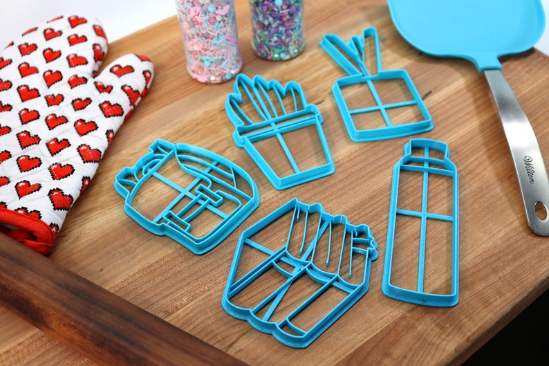K&ouml;nnte beinhalten: Ein Set mit sechs blauen Plastik-Ausstechern in Form eines Kaktus, einer Geschenkbox, einer Spr&uuml;hflasche, eines Rucksacks, einer Portion Pommes und eines Blumentopfs. Die Ausstecher liegen auf einem Holzbrett.
