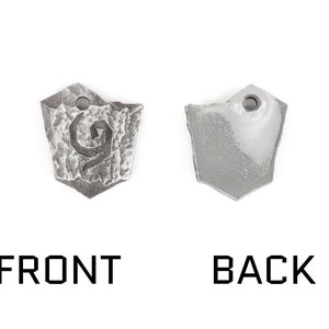May include: A silver metal pendant with a stylized design on the front and a smooth surface on the back. The pendant is shaped like a shield.