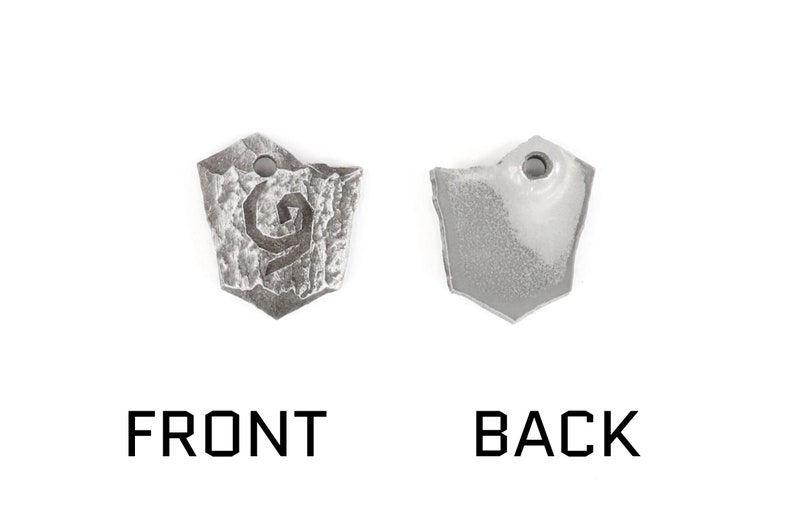 May include: A silver metal pendant with a stylized design on the front and a smooth surface on the back. The pendant is shaped like a shield.