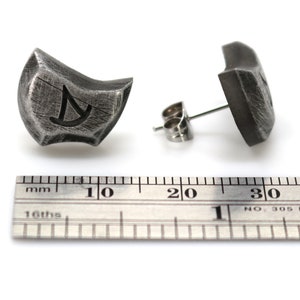 May include: A pair of silver earrings with a textured, geometric design. Each earring features a small, engraved symbol. The earrings are shown next to a ruler, measuring approximately 1 inch in length.