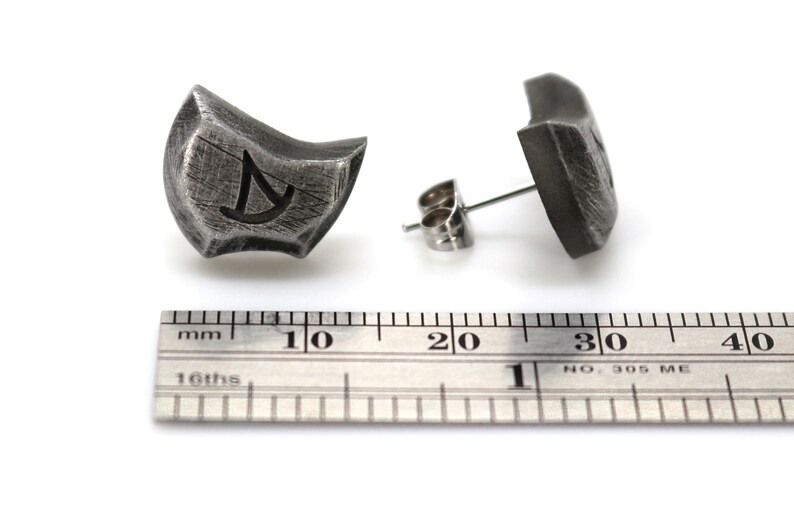 May include: A pair of silver earrings with a textured, geometric design. Each earring features a small, engraved symbol. The earrings are shown next to a ruler, measuring approximately 1 inch in length.
