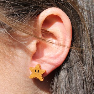 May include: Gold-colored star-shaped earrings with a pixelated design. The earrings are worn in a person's ear.