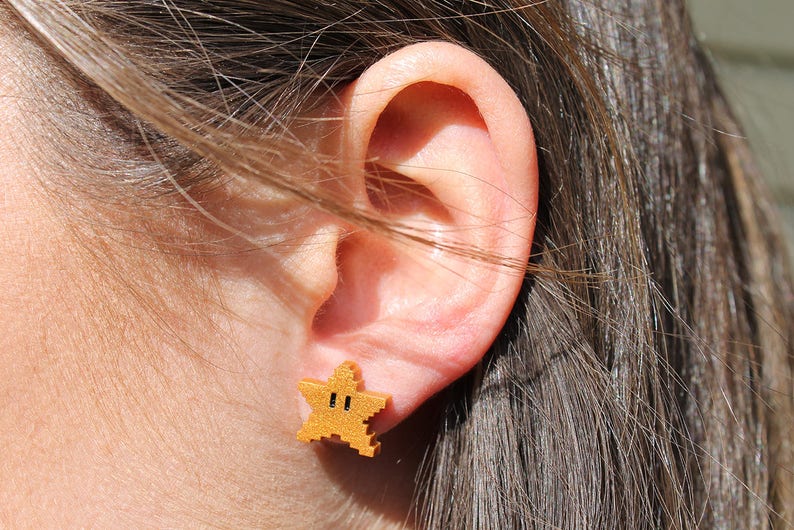 May include: Gold-colored star-shaped earrings with a pixelated design. The earrings are worn in a person's ear.