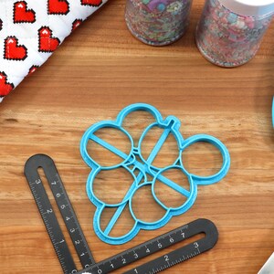 May include: Blue plastic cookie cutter shaped like a bunch of balloons. The cutter is on a wooden surface with a black ruler and a blue spatula.
