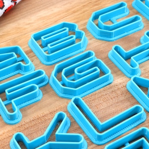 May include: A set of blue plastic alphabet cookie cutters. The cutters are arranged on a wooden surface.