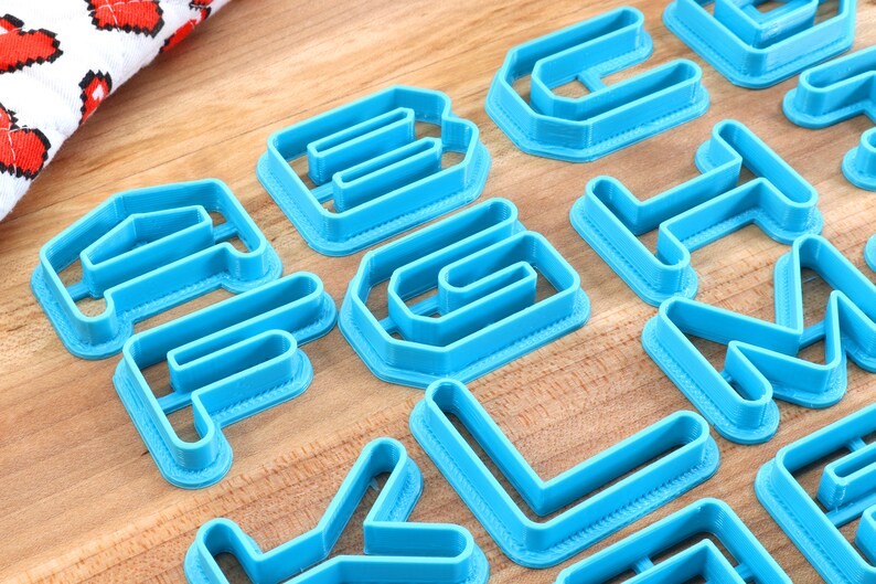 May include: A set of blue plastic alphabet cookie cutters. The cutters are arranged on a wooden surface.