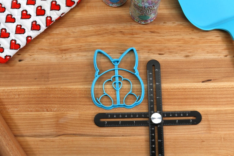 Pu&ograve; includere: Un tagliapasta in plastica blu a forma di sedere di cane. Il tagliapasta &egrave; su un righello nero con segni bianchi.