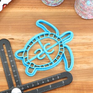 May include: Blue plastic turtle cookie cutter with a 2C design in the centre. The cutter is on a wooden surface.