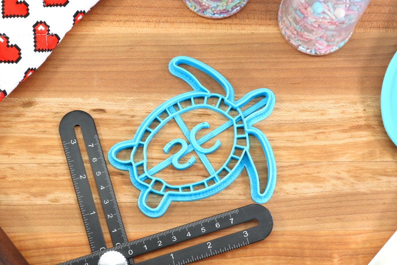 May include: Blue plastic turtle cookie cutter with a 2C design in the centre. The cutter is on a wooden surface.