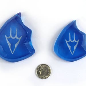 May include: Two blue glass crystal-shaped pendants with a white stylized design. The pendants are about the size of a US dime.