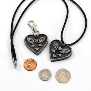 Puede incluir: Dos colgantes de plata en forma de coraz&oacute;n con un dise&ntilde;o de trifuerza alada, uno en un collar de cord&oacute;n negro y el otro en un llavero de cord&oacute;n negro. Los colgantes se muestran junto a tres monedas para comparar el tama&ntilde;o.