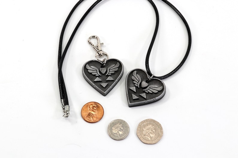 Puede incluir: Dos colgantes de plata en forma de coraz&oacute;n con un dise&ntilde;o de trifuerza alada, uno en un collar de cord&oacute;n negro y el otro en un llavero de cord&oacute;n negro. Los colgantes se muestran junto a tres monedas para comparar el tama&ntilde;o.