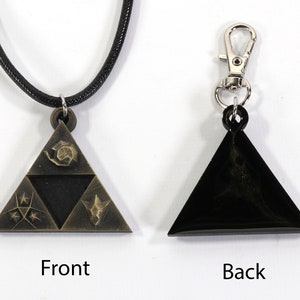 May include: A gold-colored triangular pendant with a black cord necklace. The pendant features a stylized design of a triangle with three symbols inside. The back of the pendant is black.