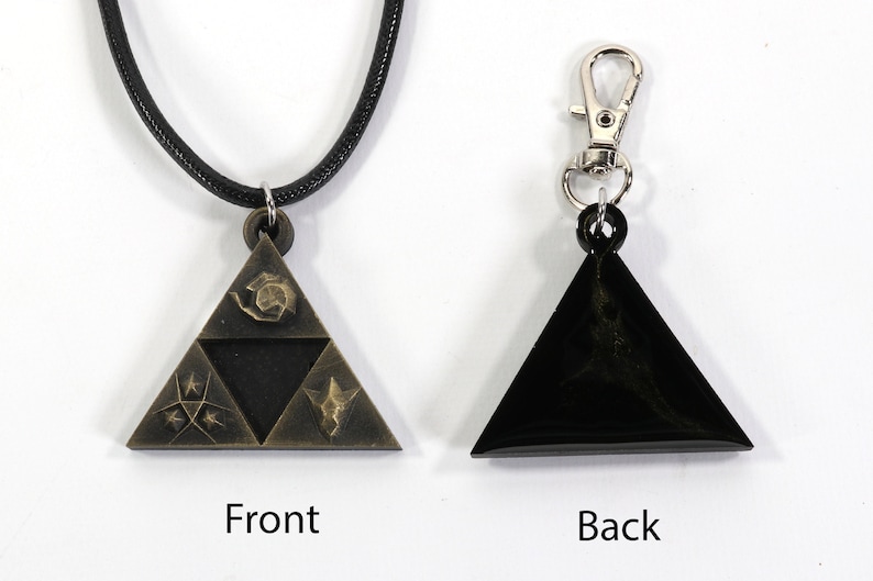 May include: A gold-colored triangular pendant with a black cord necklace. The pendant features a stylized design of a triangle with three symbols inside. The back of the pendant is black.