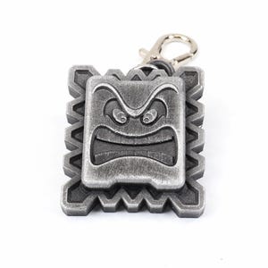 May include: A silver-colored keychain featuring a cartoon face with an angry expression. The face is surrounded by a jagged, geometric border.