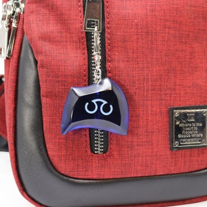 May include: A red crossbody bag with a black zipper and a blue charm with a white symbol. The bag has a black leather trim and a silver tag that reads "Where is the heart to receive goods where goods where".
