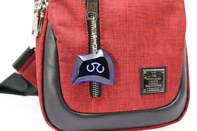 May include: A red crossbody bag with a black zipper and a blue charm with a white symbol. The bag has a black leather trim and a silver tag that reads "Where is the heart to receive goods where goods where".