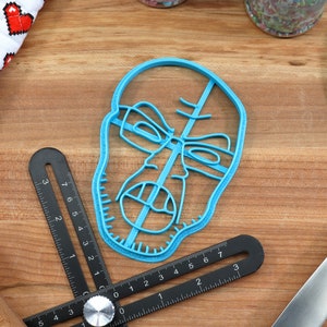 May include: A blue plastic cookie cutter shaped like a face with a serious expression. The cookie cutter is on a wooden surface with a ruler and a spatula.