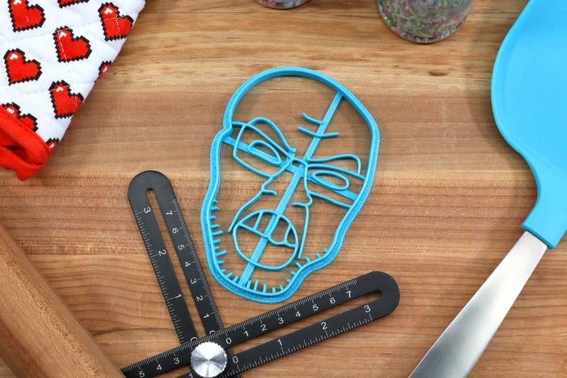 May include: A blue plastic cookie cutter shaped like a face with a serious expression. The cookie cutter is on a wooden surface with a ruler and a spatula.