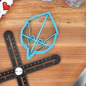 May include: A blue plastic cookie cutter shaped like a bird's head. The cutter is on a wooden surface with a black measuring tool and a blue spatula.