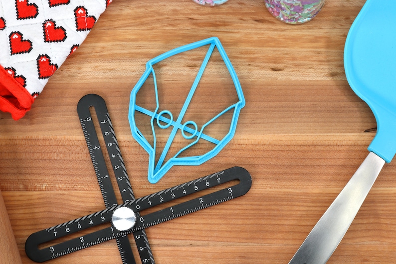 May include: A blue plastic cookie cutter shaped like a bird's head. The cutter is on a wooden surface with a black measuring tool and a blue spatula.