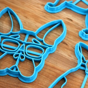 Puede incluir: Tres cortadores de galletas de pl&aacute;stico azul con forma de perro. El cortador del centro presenta un perro con gafas.